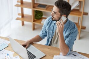 Portrait of handsome young bearded male financier talks via smart phone, tries to install new progra