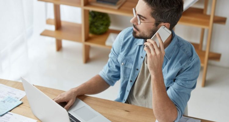 Portrait of handsome young bearded male financier talks via smart phone, tries to install new progra