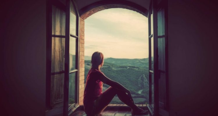 Young woman sitting in an open old window looking on the landscape of Tuscany, Italy.